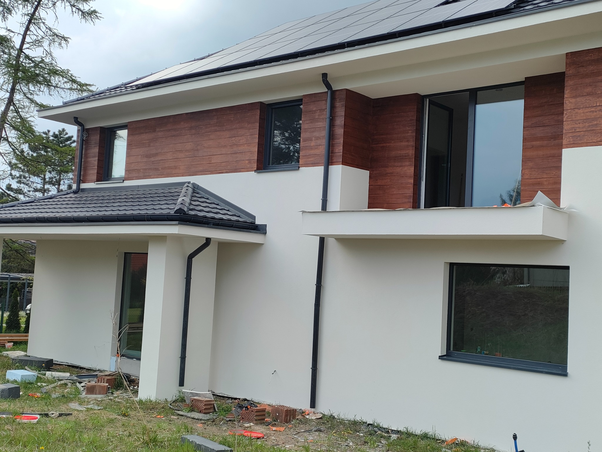 Nowoczesny dom z elewacją w kolorze bieli i drewna, okna w ciemnych ramach, panele słoneczne na dachu. Widoczne materiały budowlane.