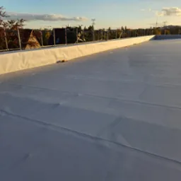 Biała, płaska powierzchnia dachu z widocznymi łączeniami, zabezpieczona barierkami, z widokiem na zabudowę miejską i zachodzące słońce.