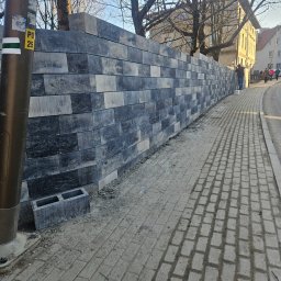 HADES MATEUSZ PIĘTA - Wzniesiona ściana z szarych bloczków betonowych wzdłuż chodnika i ulicy, widoczny słup oświetleniowy z naklejką 'PO 25' oraz pojedynczy bloczek obok, na drugim planie przechodnie i budynki.