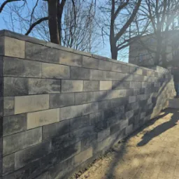 Wysoki mur z szarych, betonowych bloków o zróżnicowanej kolorystyce, zakończony elementami wieńczącymi, z widocznymi osobami w tle i drzewami bez liści na górze zdjęcia.