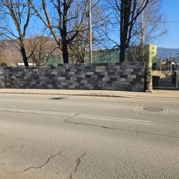 HADES MATEUSZ PIĘTA - Nowo wymurowany, wysoki mur z szaro-czarnej cegły klinkierowej, widoczni dwaj murarze na rusztowaniu, w tle drzewa bez liści i budynek mieszkalny.