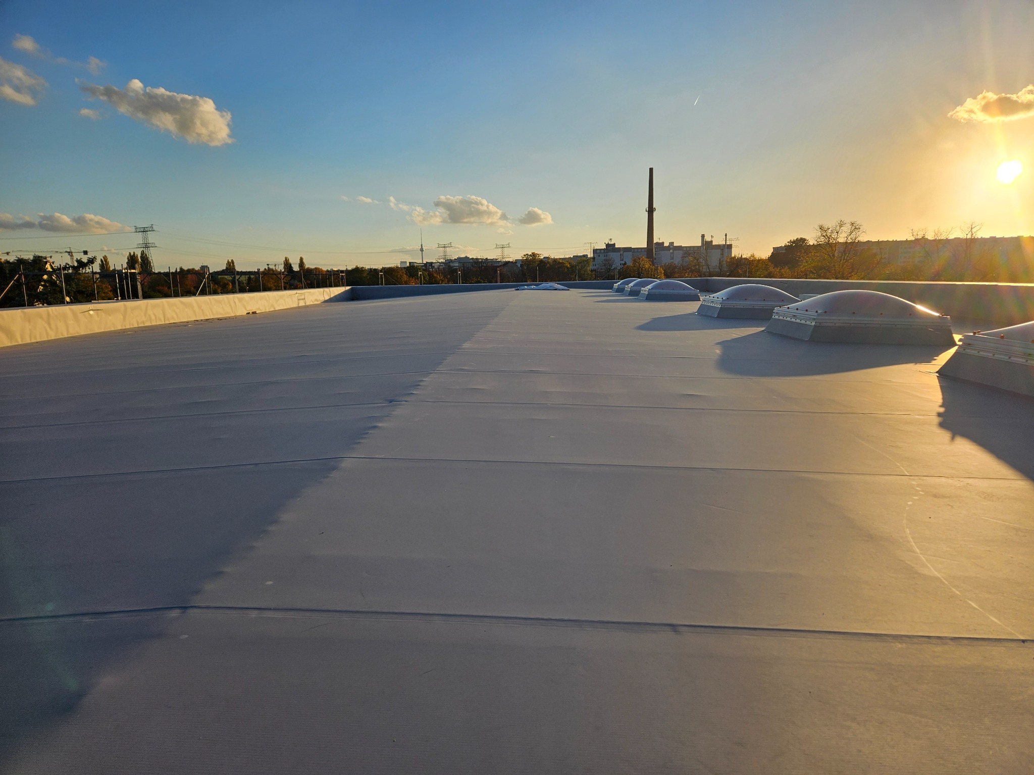 Szary dach płaski pokryty membraną hydroizolacyjną z świetlikami kopułowymi, widok z góry, w tle zabudowania miejskie i zachodzące słońce.