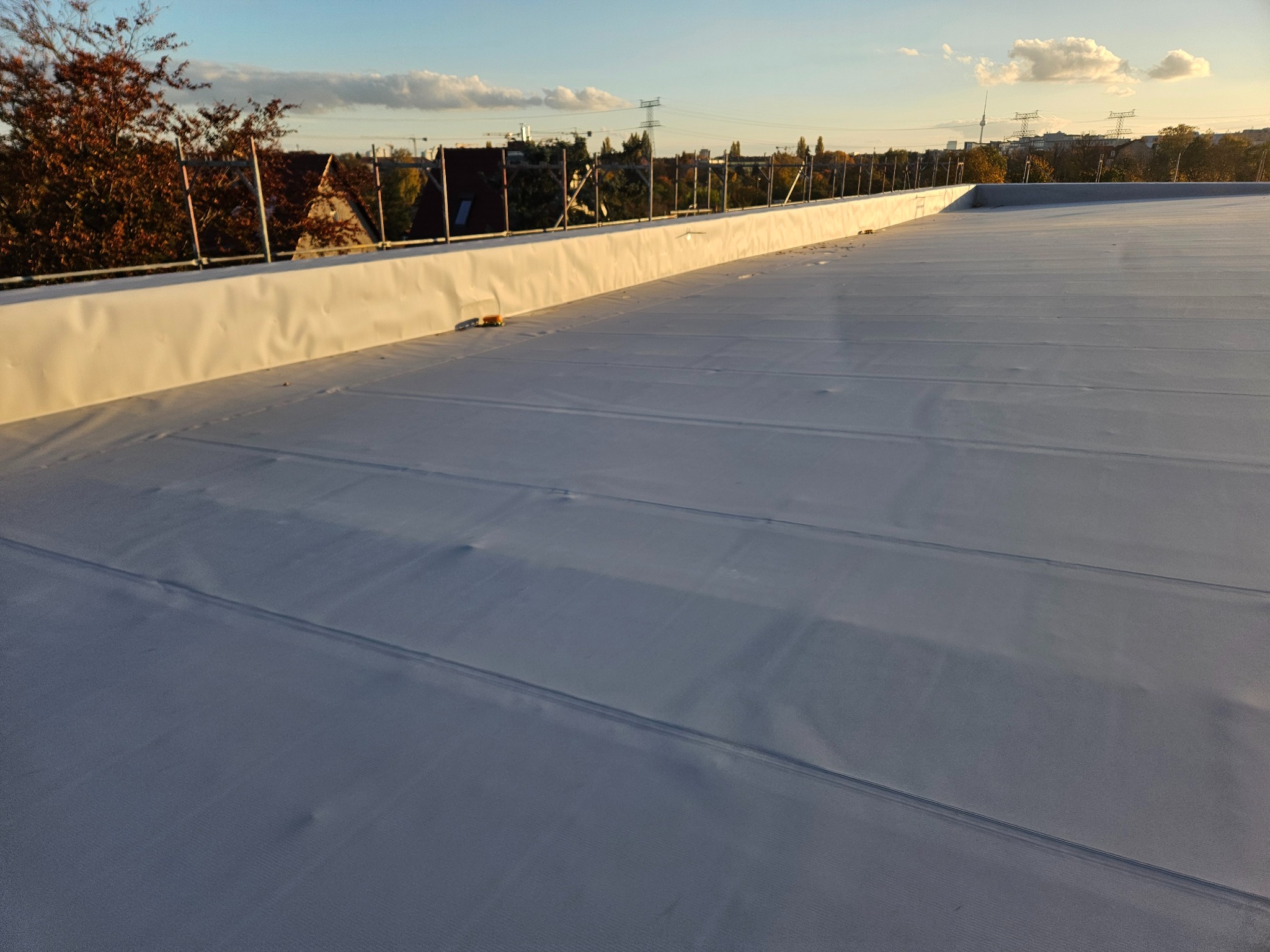 Biała, płaska powierzchnia dachu z widocznymi łączeniami, zabezpieczona barierkami, z widokiem na zabudowę miejską i zachodzące słońce.