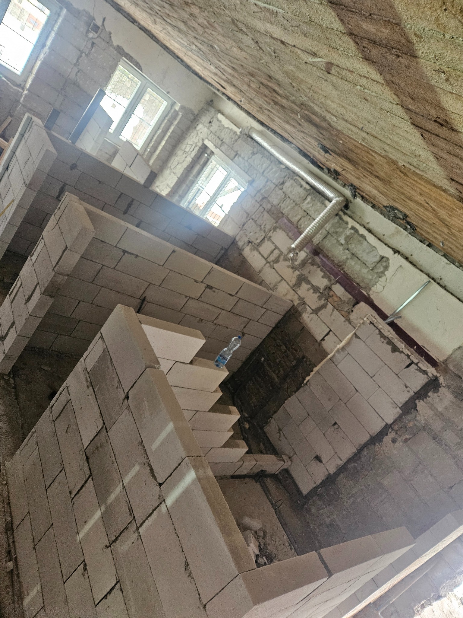 Wnętrze w trakcie budowy z widocznymi ścianami z białych bloczków, drewnianym stropem i fragmentami instalacji wentylacyjnej, butelka wody na murze.