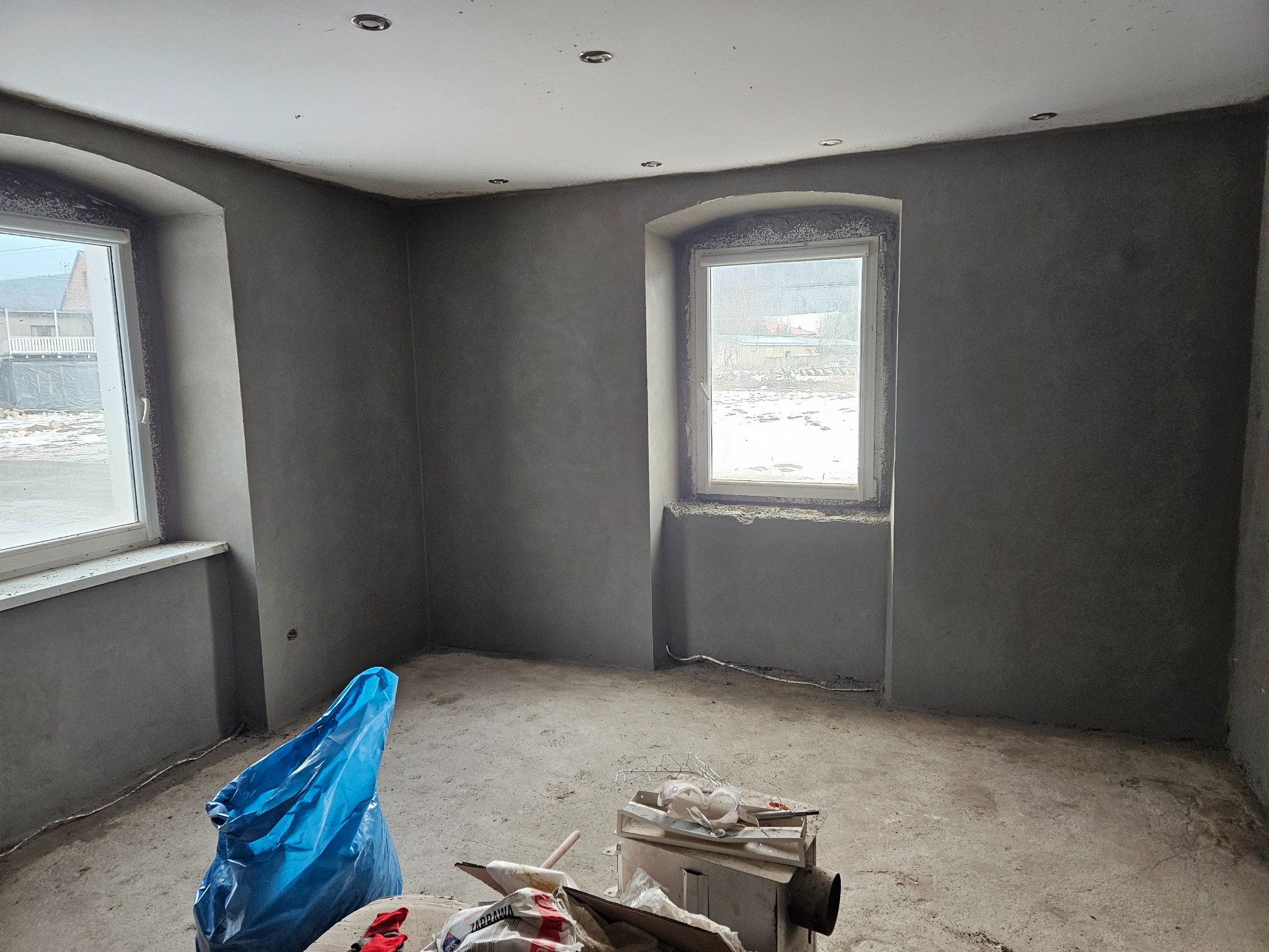 Surowe, szare wnętrze w trakcie prac wykończeniowych: świeżo otynkowane ściany z dwoma oknami o zaokrąglonych nadprożach, niebieski worek na gruz i narzędzia na podłodze.