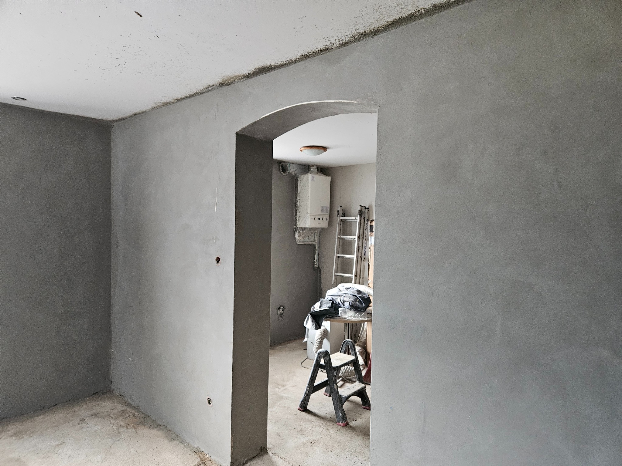 Surowe, szare ściany z widocznym wejściem łukowym do pomieszczenia z piecem gazowym i rozstawioną drabiną, przygotowane do dalszych prac wykończeniowych.