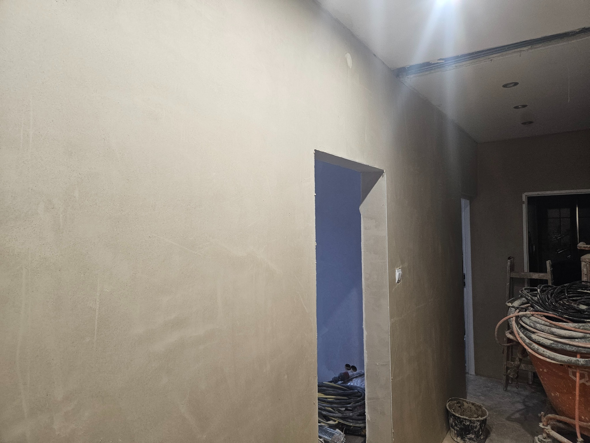 Wnętrze w trakcie prac wykończeniowych: świeżo otynkowana ściana w odcieniach beżu, widoczne przewody elektryczne i narzędzia w otworze drzwiowym oraz sprzęt do tynkowania w tle.