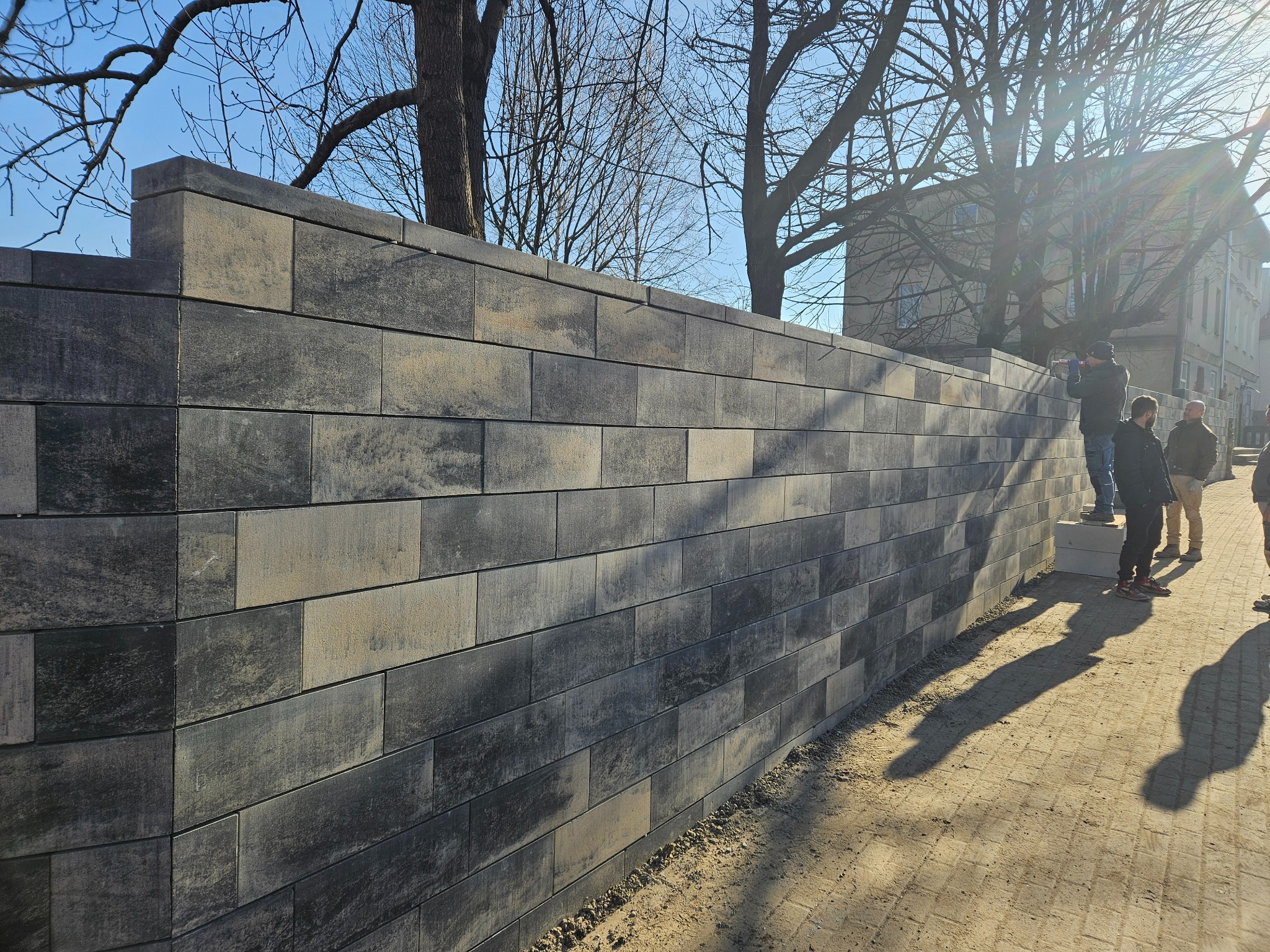 Wysoki mur z szarych, betonowych bloków o zróżnicowanej kolorystyce, zakończony elementami wieńczącymi, z widocznymi osobami w tle i drzewami bez liści na górze zdjęcia.