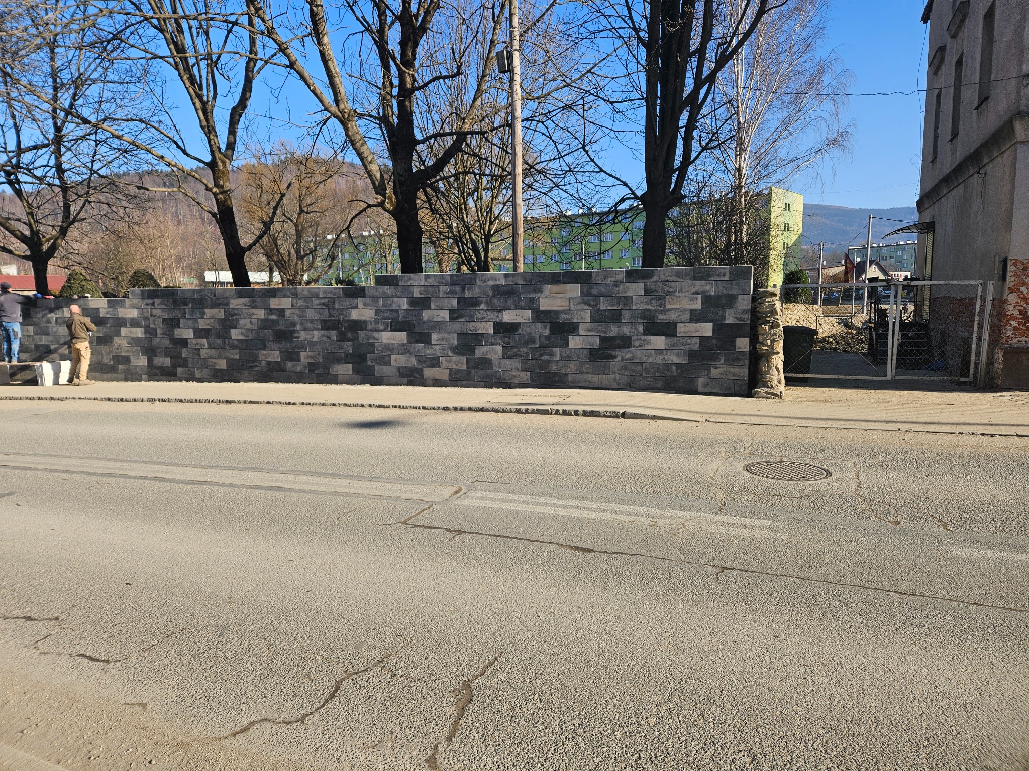 Nowo wymurowany, wysoki mur z szaro-czarnej cegły klinkierowej, widoczni dwaj murarze na rusztowaniu, w tle drzewa bez liści i budynek mieszkalny.
