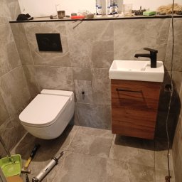 AMKORBUD - Zawieszona toaleta i umywalka z szafką w trakcie montażu w łazience wykończonej szarymi płytkami; widoczne narzędzia i materiały budowlane.