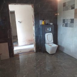 AMKORBUD - Remont łazienki w trakcie: położona szara marmurowa podłoga, częściowo wykończone ściany z płytek w odcieniach szarości, widoczny stelaż podtynkowy WC z otwartą klapą, niedokończone obramowanie drzwi.