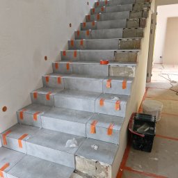 AMKORBUD - Montaż szarych płytek na schodach w trakcie remontu, zabezpieczone pomarańczową taśmą, widoczny surowy beton i cegła, na podłodze wiadro i pojemnik z narzędziami.