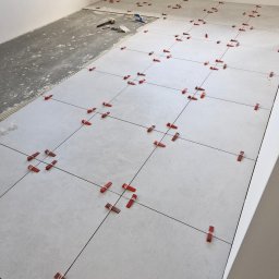 AMKORBUD - Układanie jasnych, kwadratowych płytek podłogowych z użyciem czerwonych klipsów poziomujących, widoczny fragment surowej posadzki i narzędzia glazurnicze w rogu pomieszczenia.