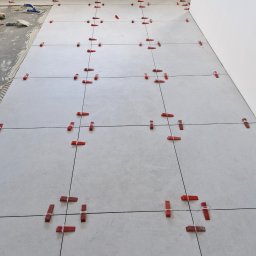 AMKORBUD - Układanie jasnych, kwadratowych płytek podłogowych z użyciem czerwonych klinów i poziomowania, widoczny fragment przygotowanego podłoża z klejem.