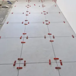 Układanie jasnych, kwadratowych płytek podłogowych z użyciem czerwonych klinów i poziomowania, widoczny fragment przygotowanego podłoża z klejem.