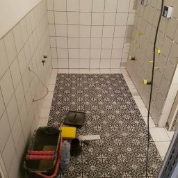 AMKORBUD - Wnętrze łazienki w trakcie remontu: ułożona wzorzysta podłoga, częściowo wykafelkowane ściany z widocznymi uchwytami do armatury, obok wiadro z narzędziami do malowania.