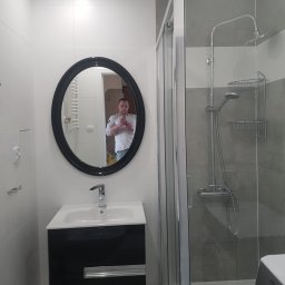 AMKORBUD - Wyremontowana łazienka z kabiną prysznicową, umywalką z czarną szafką i okrągłym lustrem, w którym odbija się osoba robiąca zdjęcie telefonem.