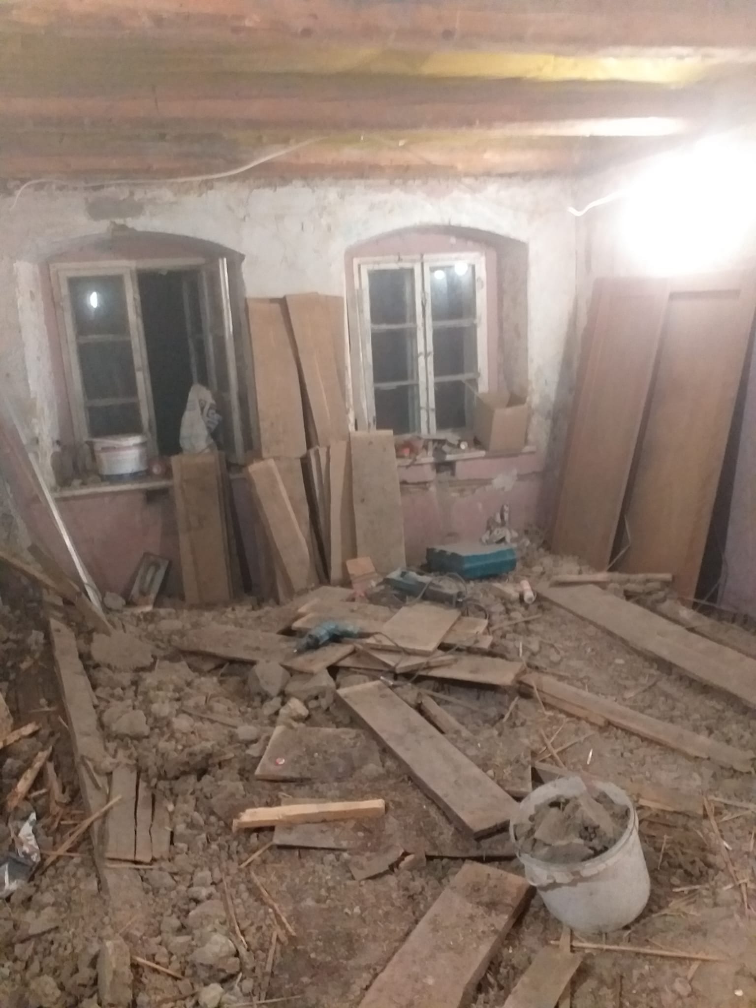 Wnętrze pokoju w trakcie prac remontowych: gruz na podłodze, deski opierają się o ścianę, widoczne okna z białymi ramami, wiadro z resztkami materiałów budowlanych, narzędzia leżą na parapecie.