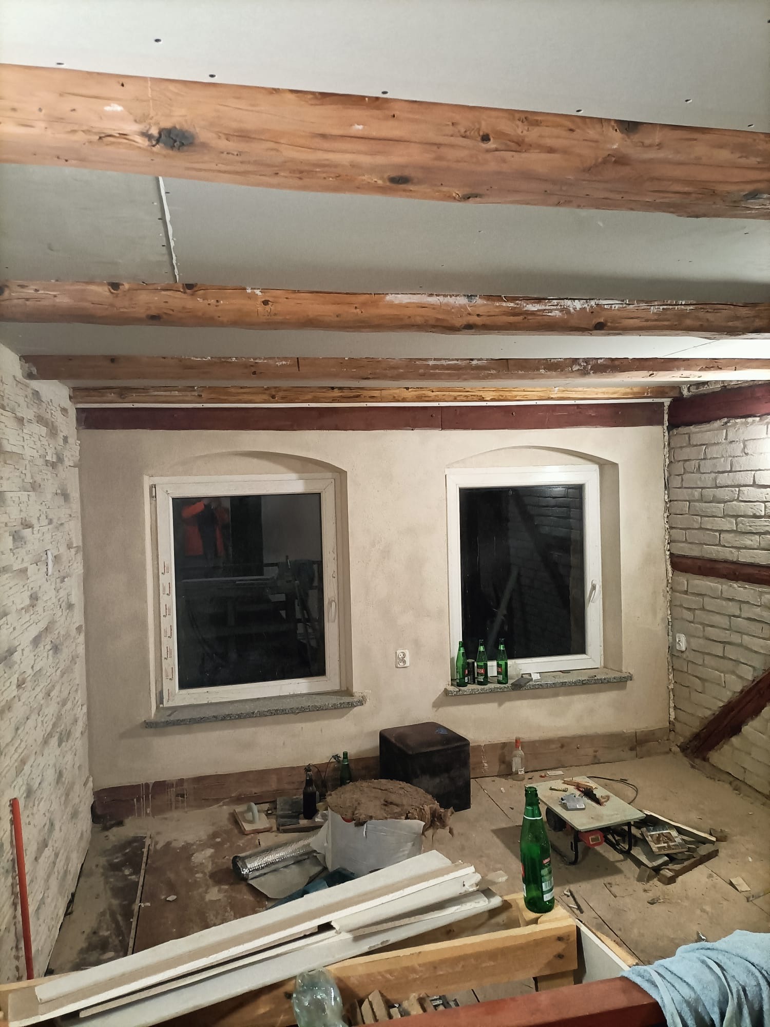 Wnętrze w trakcie prac remontowych: odsłonięte drewniane belki stropowe, ściany częściowo otynkowane i wykończone imitacją cegły, okna z butelkami na parapecie, widoczne materiały budowlane...