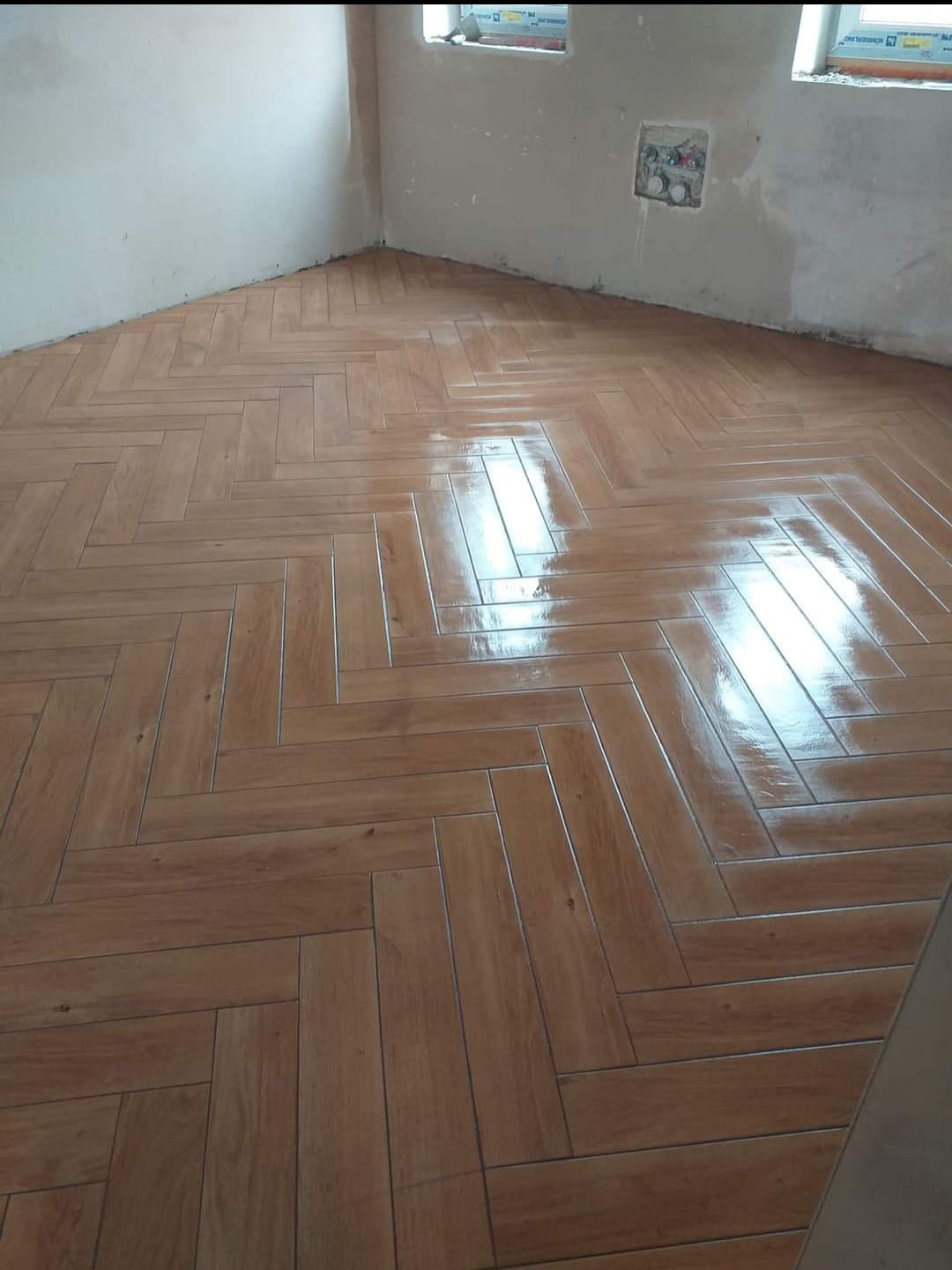 Ułożona w jodełkę, błyszcząca, brązowa podłoga z płytek imitujących drewno w trakcie remontu łazienki, widoczne okna z folią ochronną i surowe ściany.