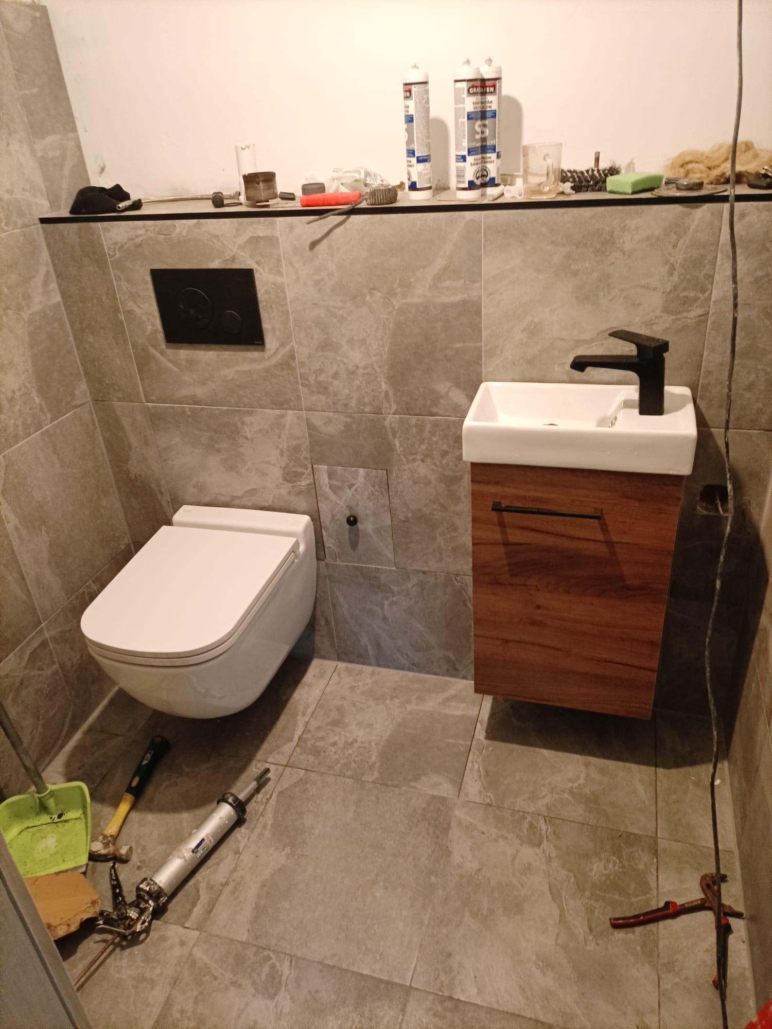 Zawieszona toaleta i umywalka z szafką w trakcie montażu w łazience wykończonej szarymi płytkami; widoczne narzędzia i materiały budowlane.