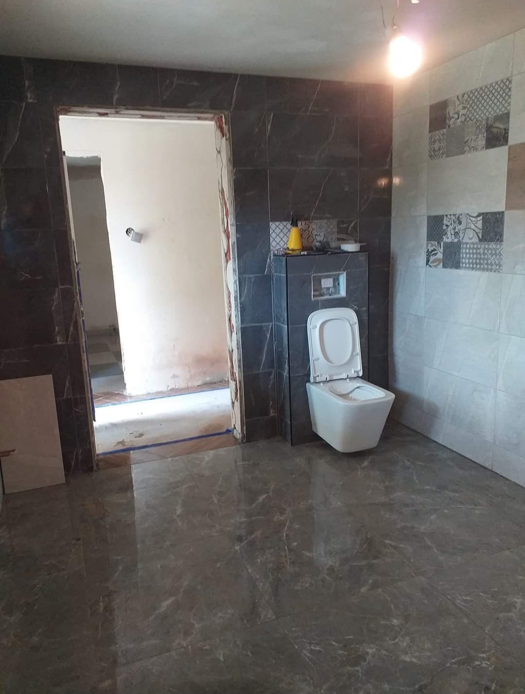 Remont łazienki w trakcie: położona szara marmurowa podłoga, częściowo wykończone ściany z płytek w odcieniach szarości, widoczny stelaż podtynkowy WC z otwartą klapą, niedokończone obramowanie drzwi.