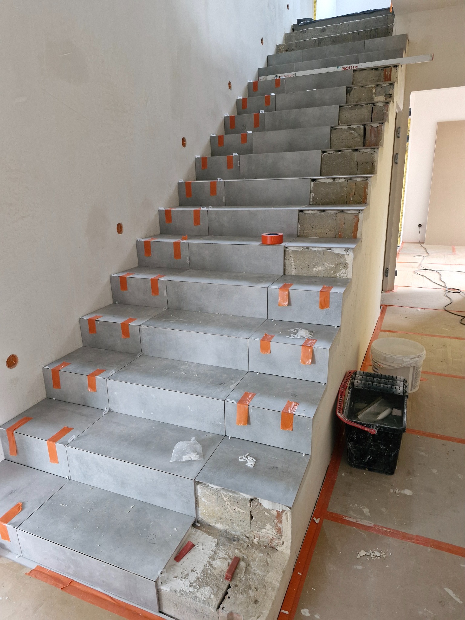 Montaż szarych płytek na schodach w trakcie remontu, zabezpieczone pomarańczową taśmą, widoczny surowy beton i cegła, na podłodze wiadro i pojemnik z narzędziami.