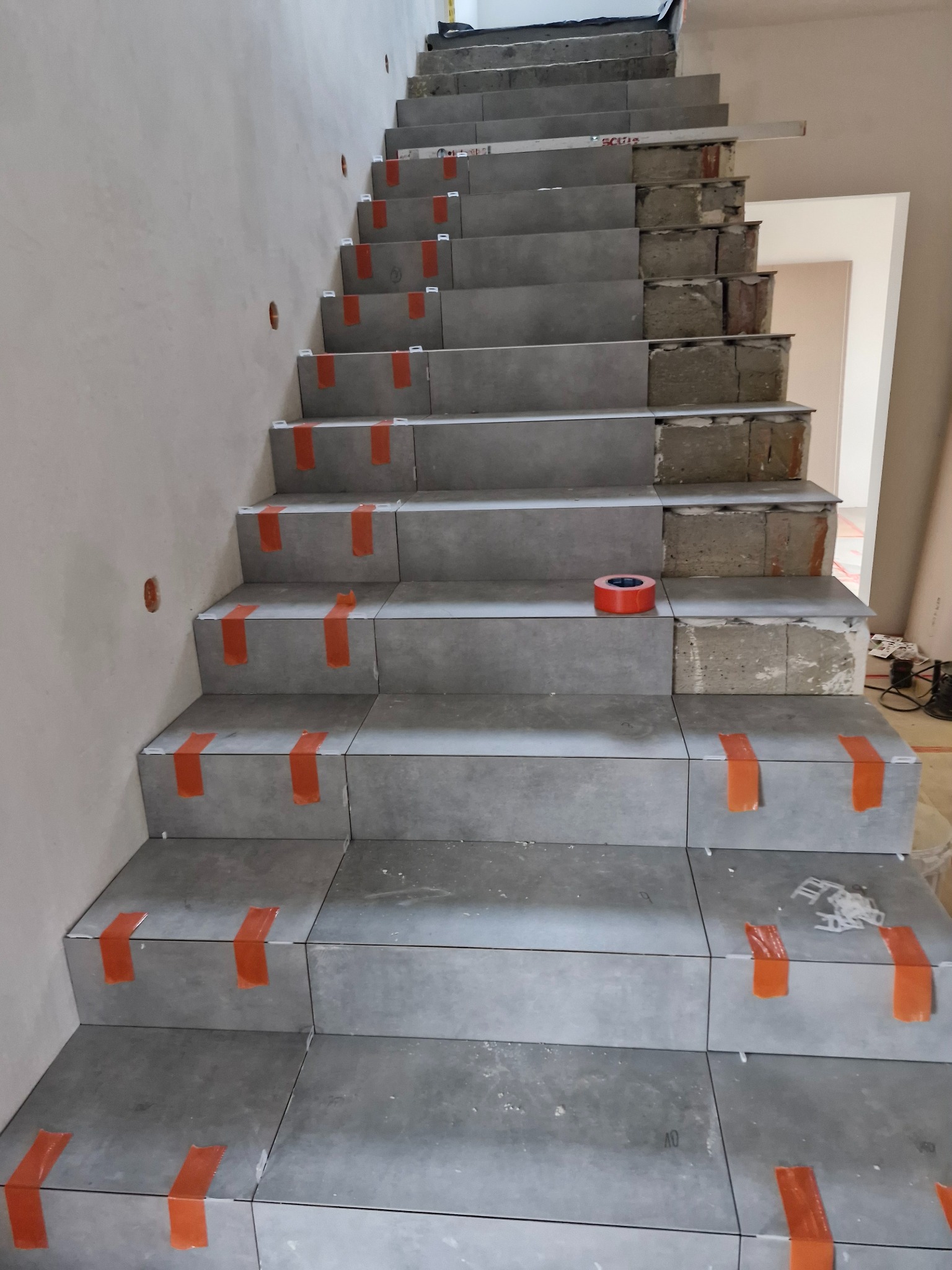 Montaż szarych płytek na betonowych schodach w trakcie prac wykończeniowych, zabezpieczonych pomarańczową taśmą, widoczna poziomica i resztki kleju.