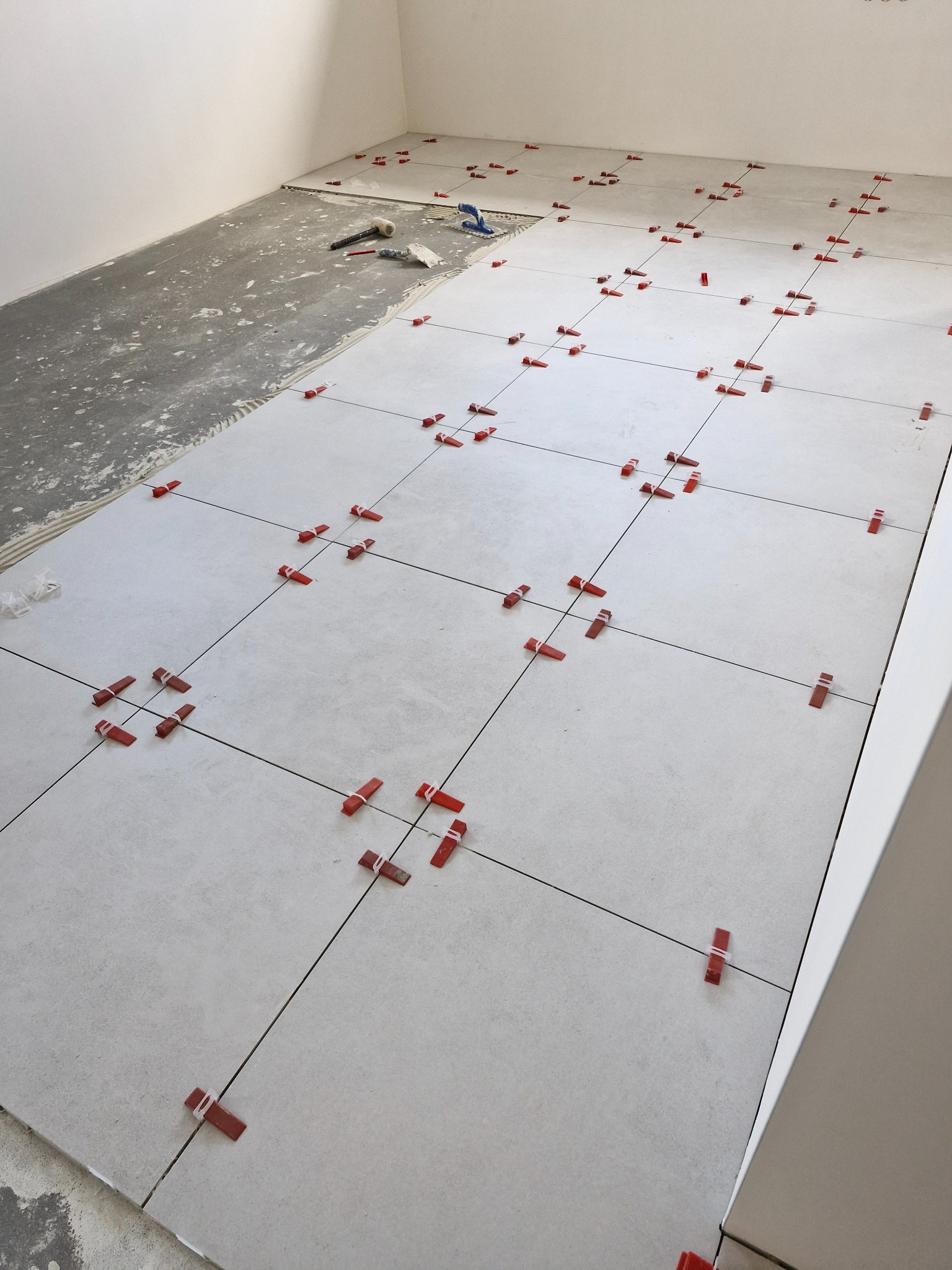 Układanie jasnych, kwadratowych płytek podłogowych z użyciem czerwonych klipsów poziomujących, widoczny fragment surowej posadzki i narzędzia glazurnicze w rogu pomieszczenia.
