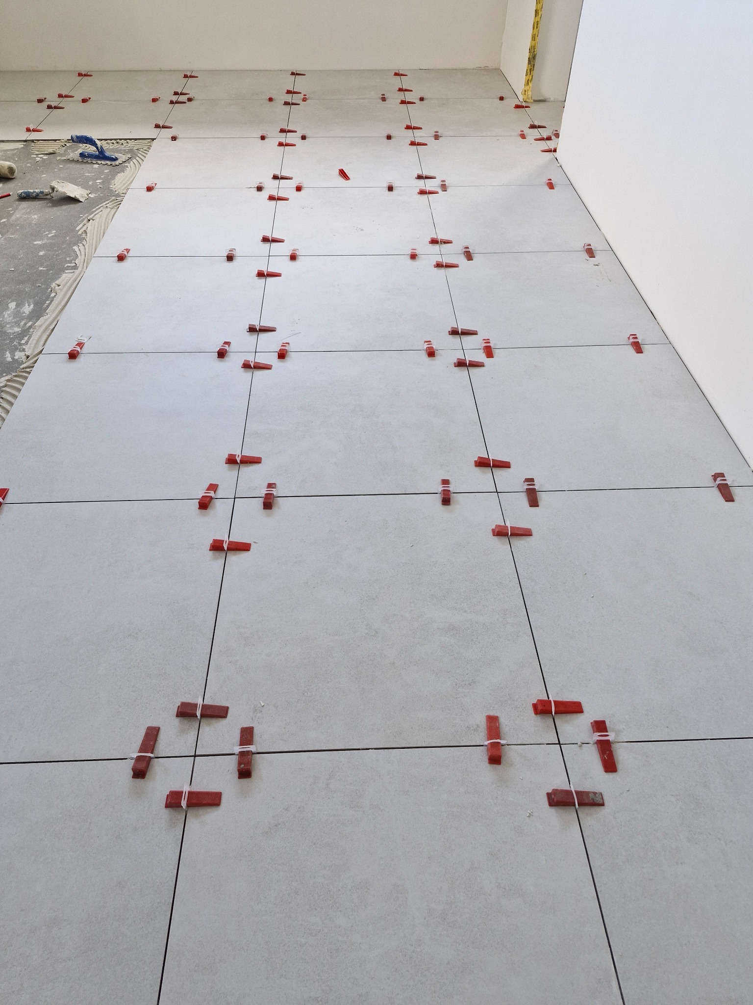 Układanie jasnych, kwadratowych płytek podłogowych z użyciem czerwonych klinów i poziomowania, widoczny fragment przygotowanego podłoża z klejem.