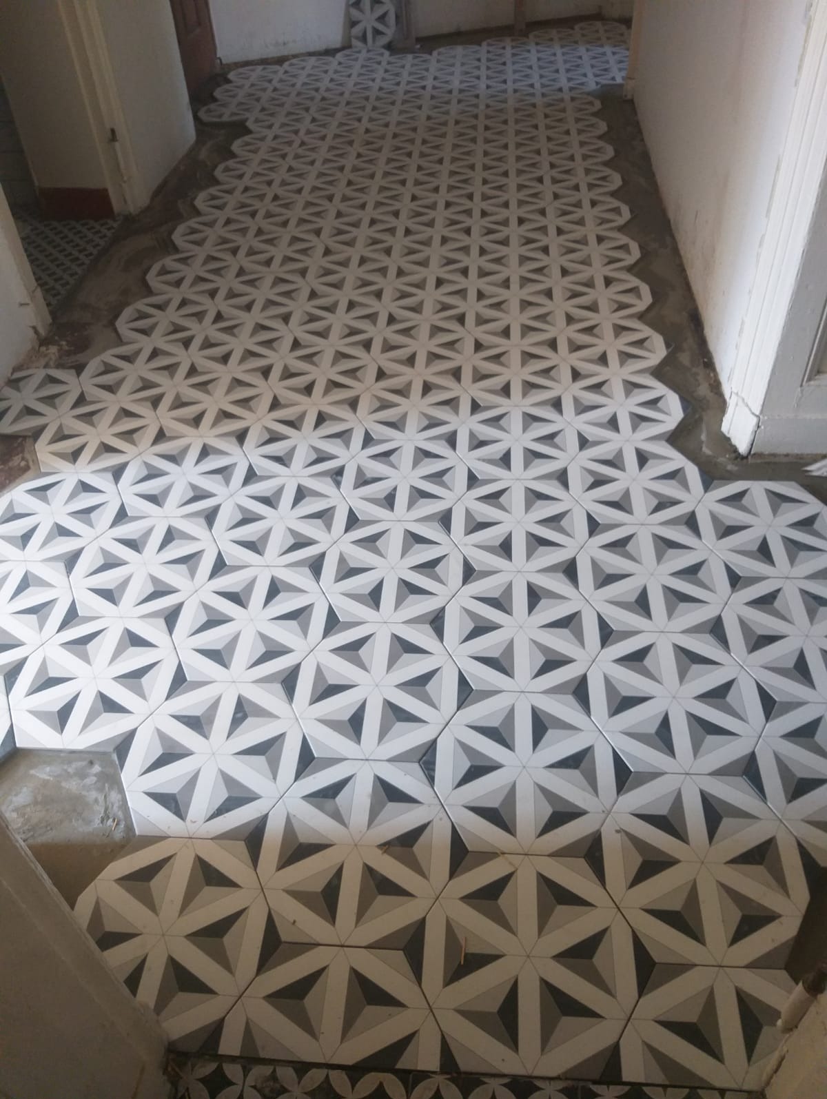 Układanie wzorzystych, heksagonalnych płytek podłogowych w odcieniach szarości i bieli, widoczny świeży cement między płytkami, częściowo ułożona podłoga w pomieszczeniu.