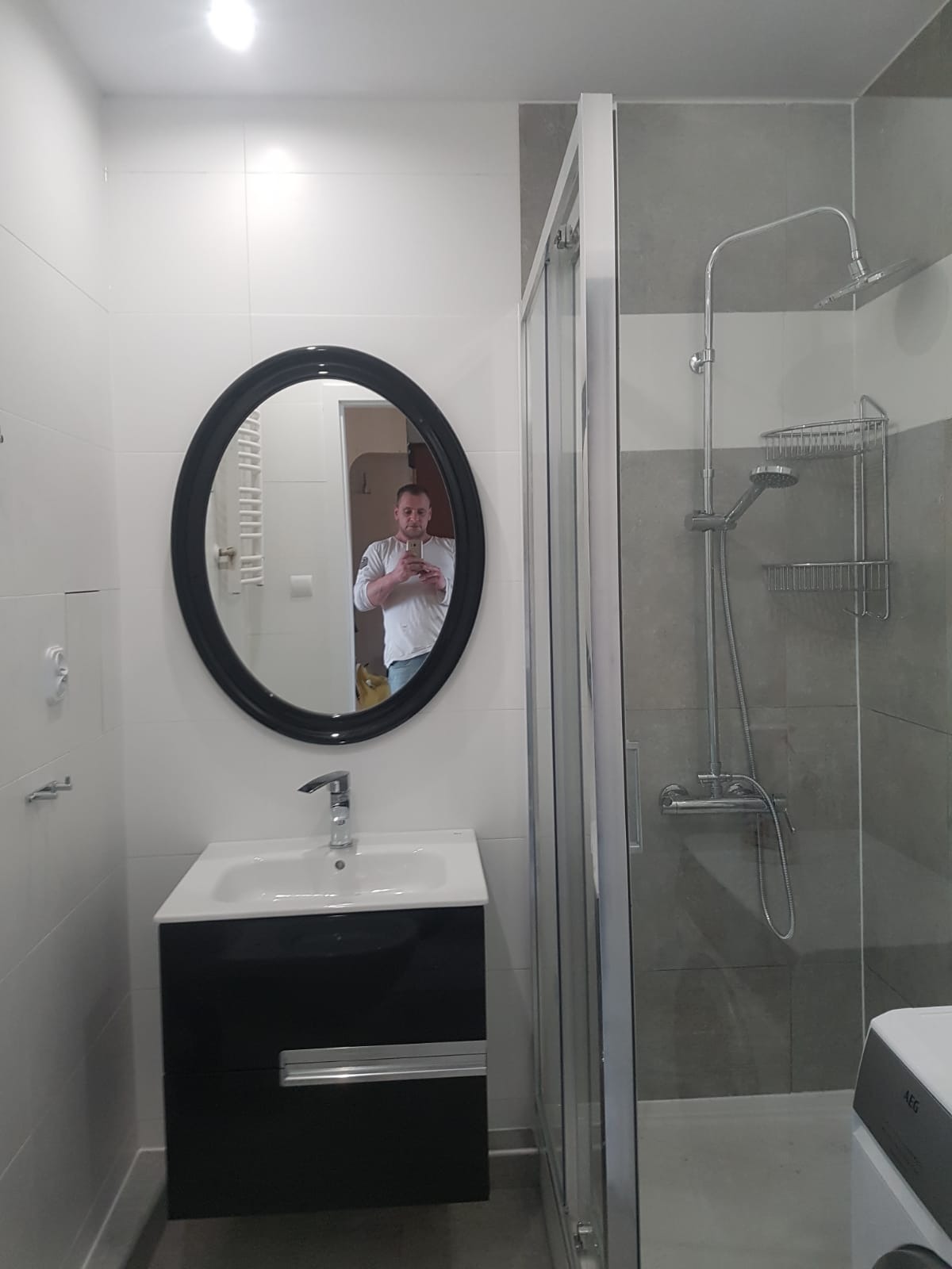 Wyremontowana łazienka z kabiną prysznicową, umywalką z czarną szafką i okrągłym lustrem, w którym odbija się osoba robiąca zdjęcie telefonem.