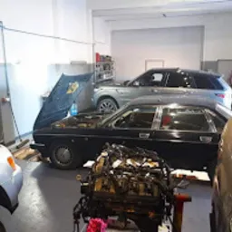 Wnętrze warsztatu samochodowego z podniesioną maską czarnego Jaguara, widoczny silnik na podłodze, w tle srebrny Range Rover i inne pojazdy.