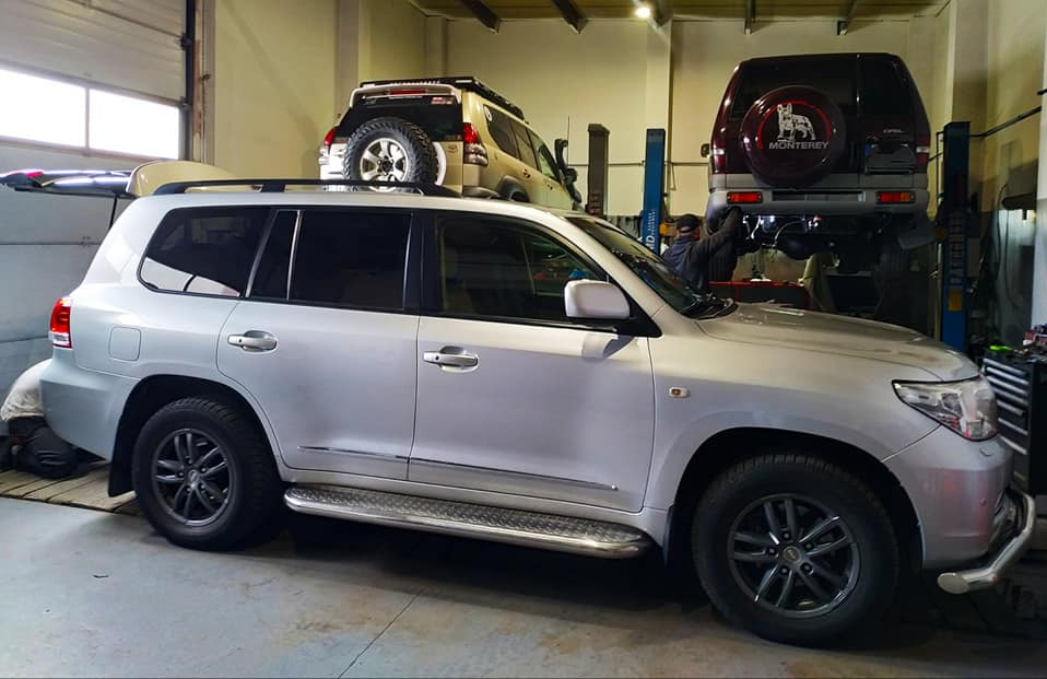 Srebrny SUV z relingami dachowymi w warsztacie; dwa inne auta na podnośnikach w tle, w tym jeden z kołem zapasowym na tylnej klapie i napisem 'Monterey'.