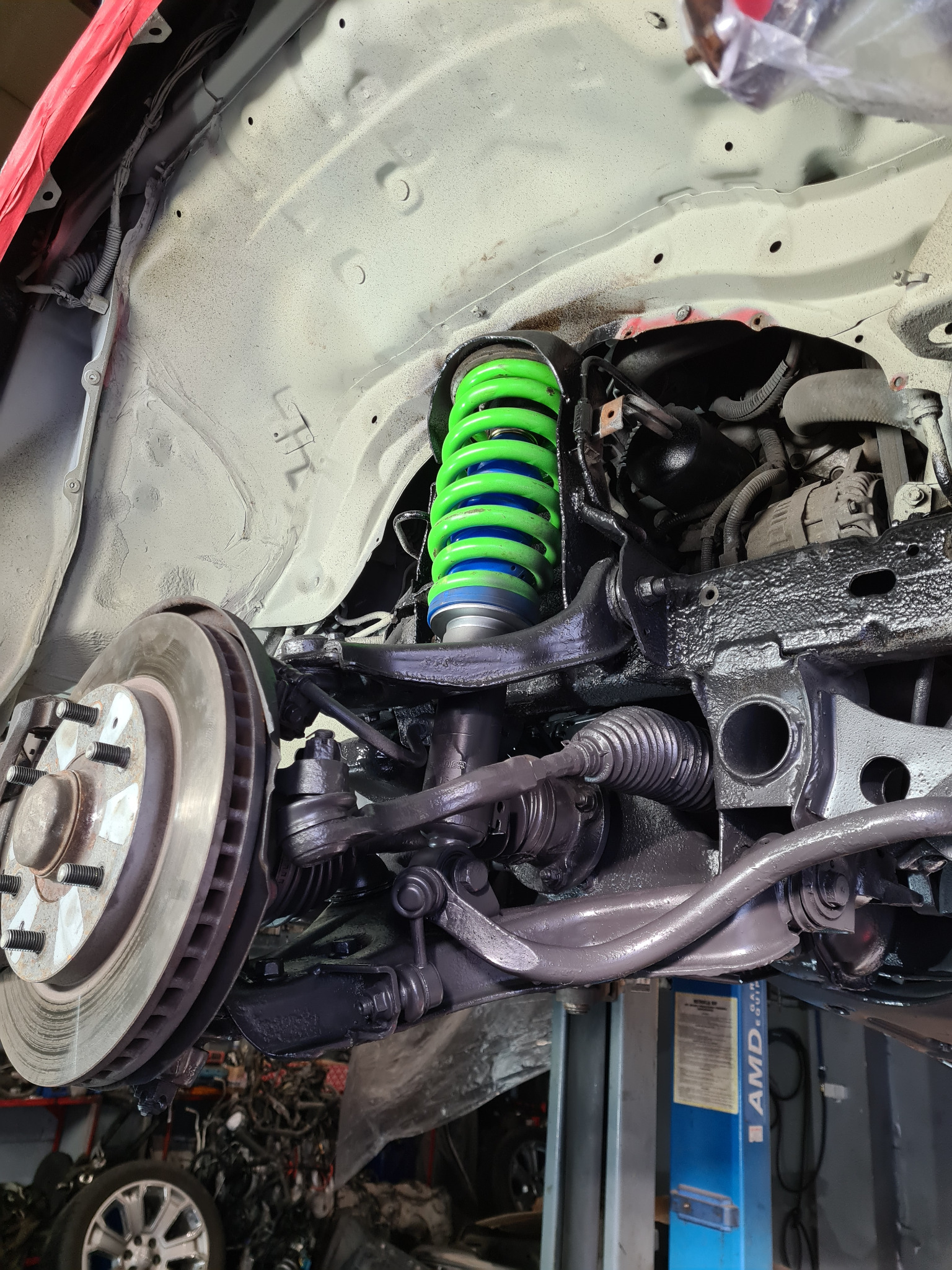 Podwozie samochodu na podnośniku z elementami zawieszenia pomalowanymi na czarno, z nową zielono-niebieską sprężyną amortyzatora i tarczą hamulcową z widocznymi śrubami.