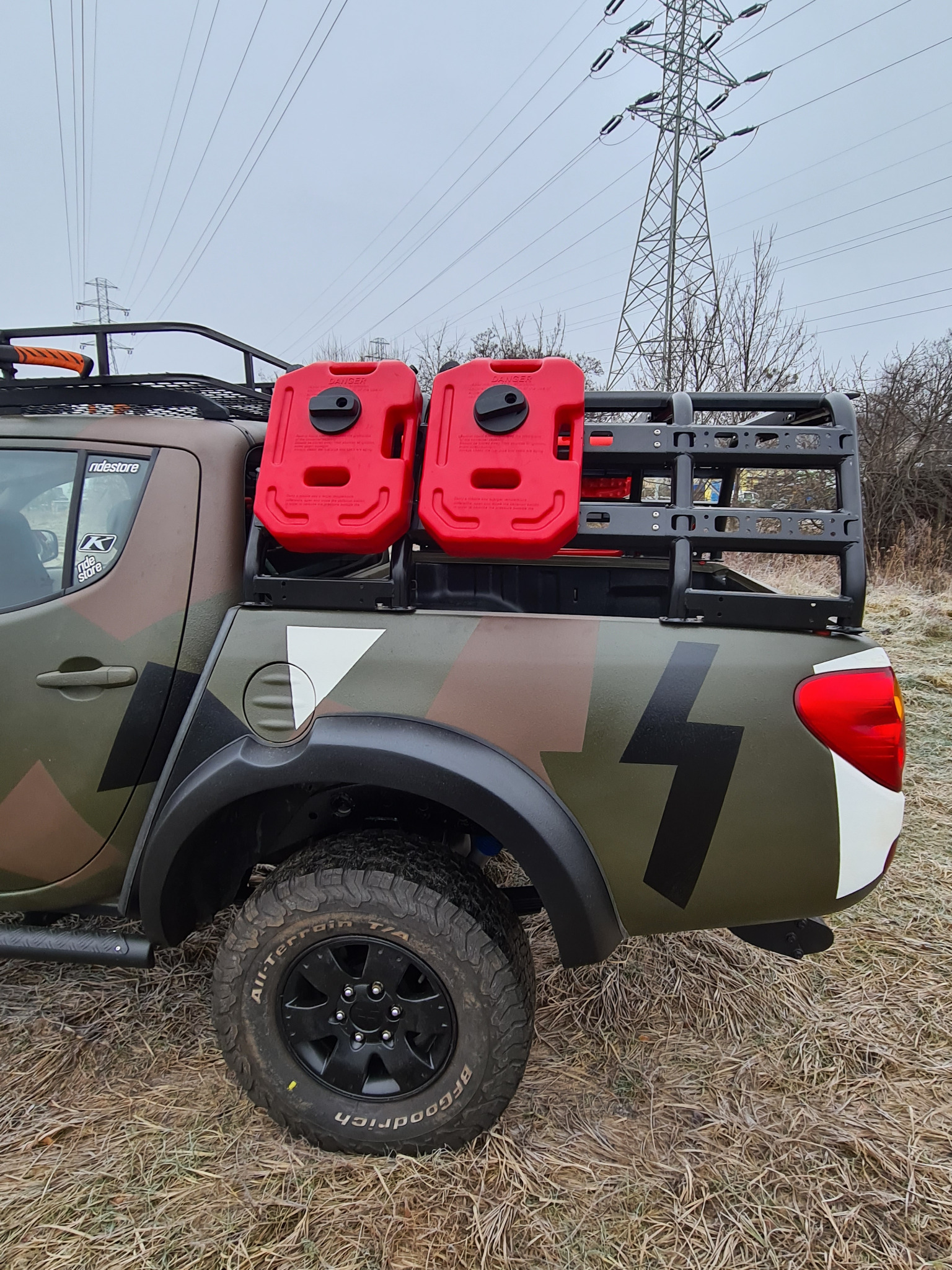 Pickup w kamuflażu z dwoma czerwonymi kanistrami na platformie bagażowej, terenowe opony i czarne felgi, słup wysokiego napięcia w tle.