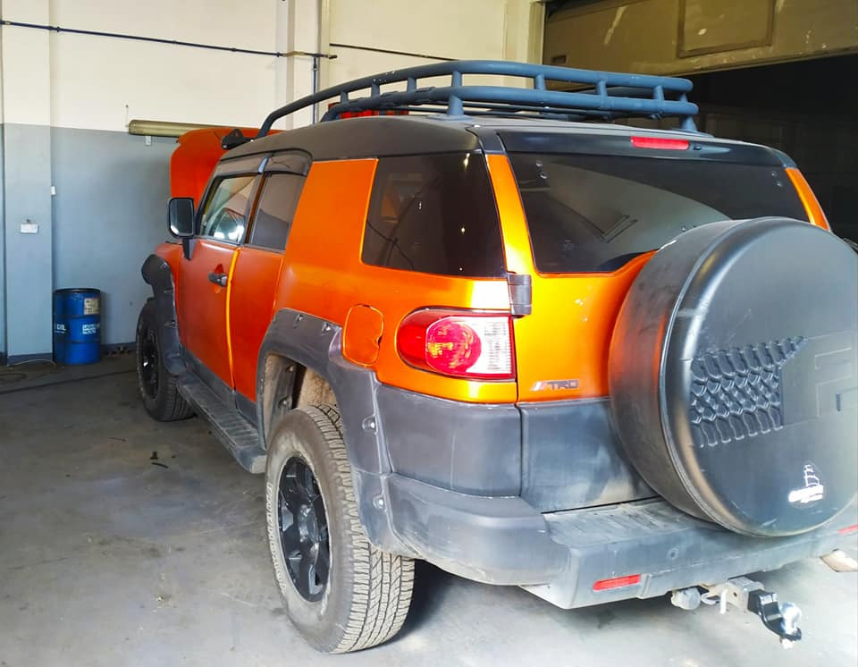 Pomarańczowy Toyota FJ Cruiser z otwartą maską w warsztacie, widoczny bagażnik dachowy i koło zapasowe z tyłu.