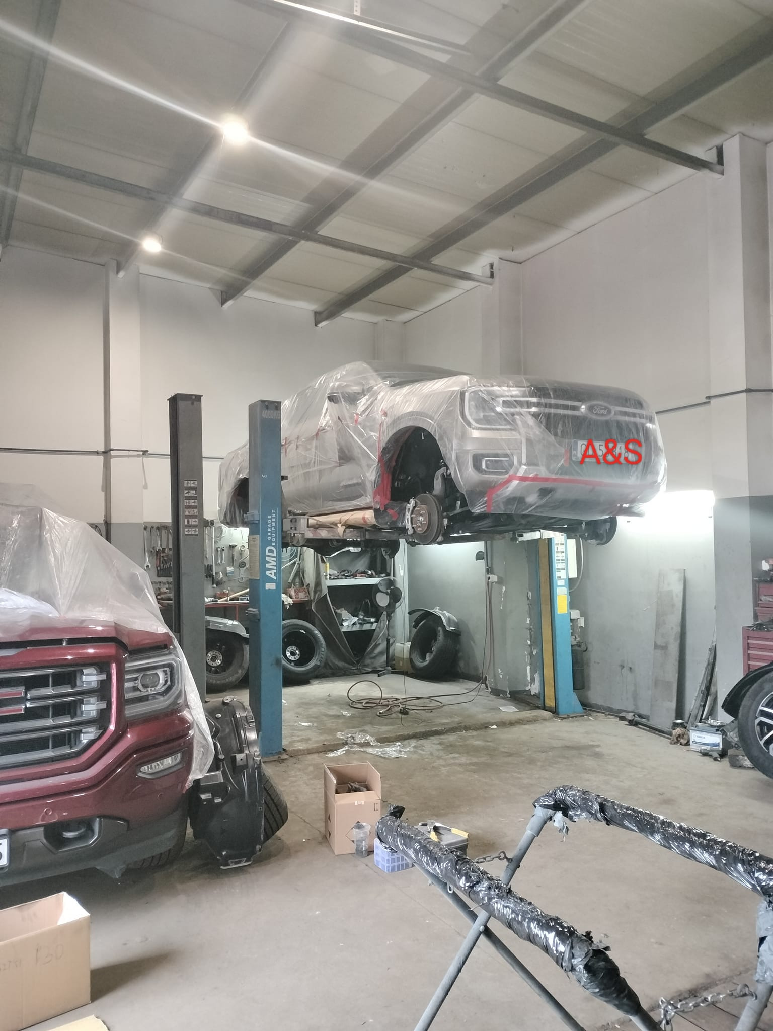 Samochód Ford Explorer na podnośniku w warsztacie, zabezpieczony folią ochronną, widoczne zdjęte koła, w tle drugi samochód przykryty folią, oświetlenie sufitowe.