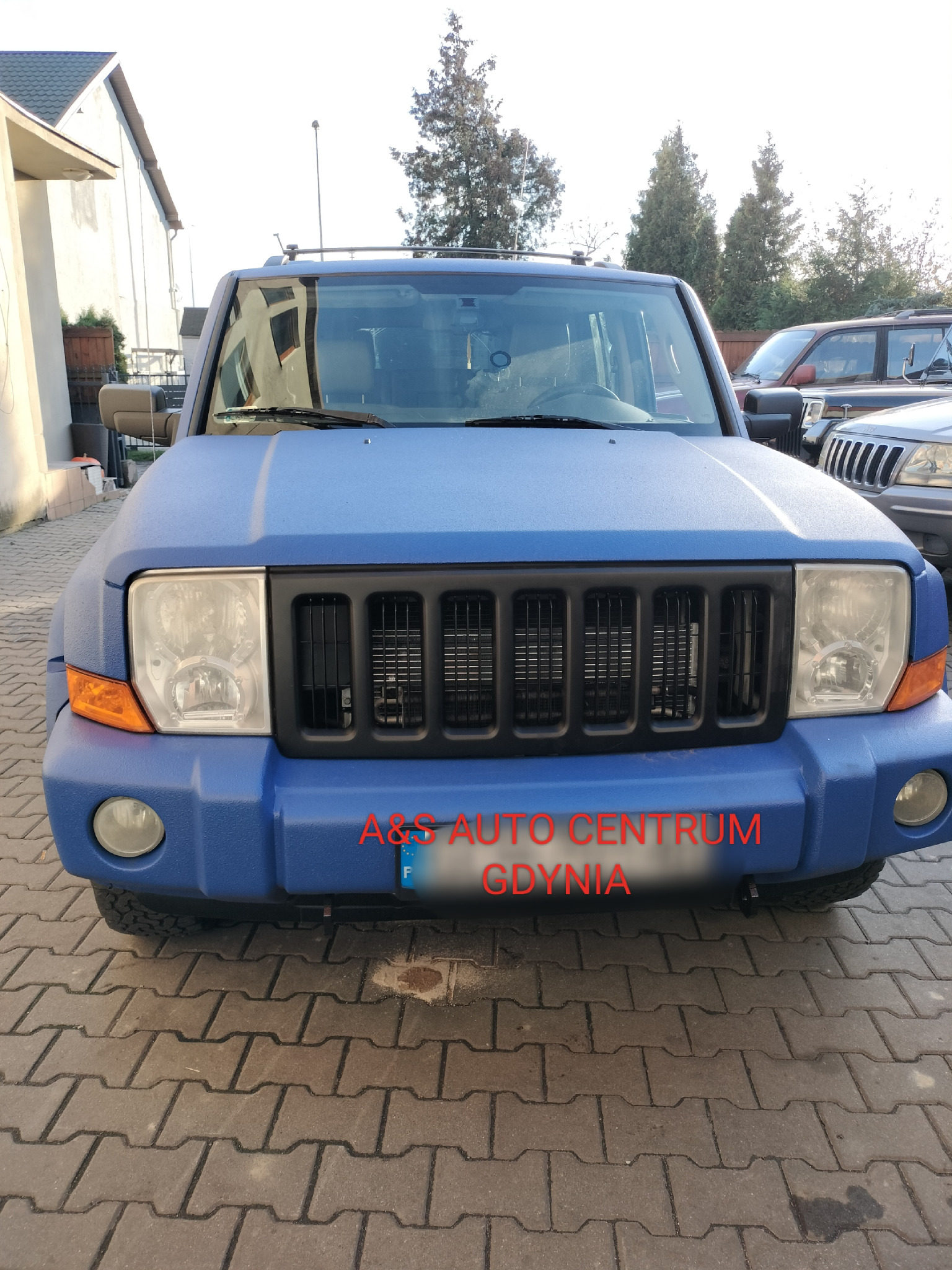 Niebieski Jeep Commander z czarną atrapą chłodnicy, pokryty matową powłoką ochronną, zaparkowany na brukowanej nawierzchni przed budynkiem.