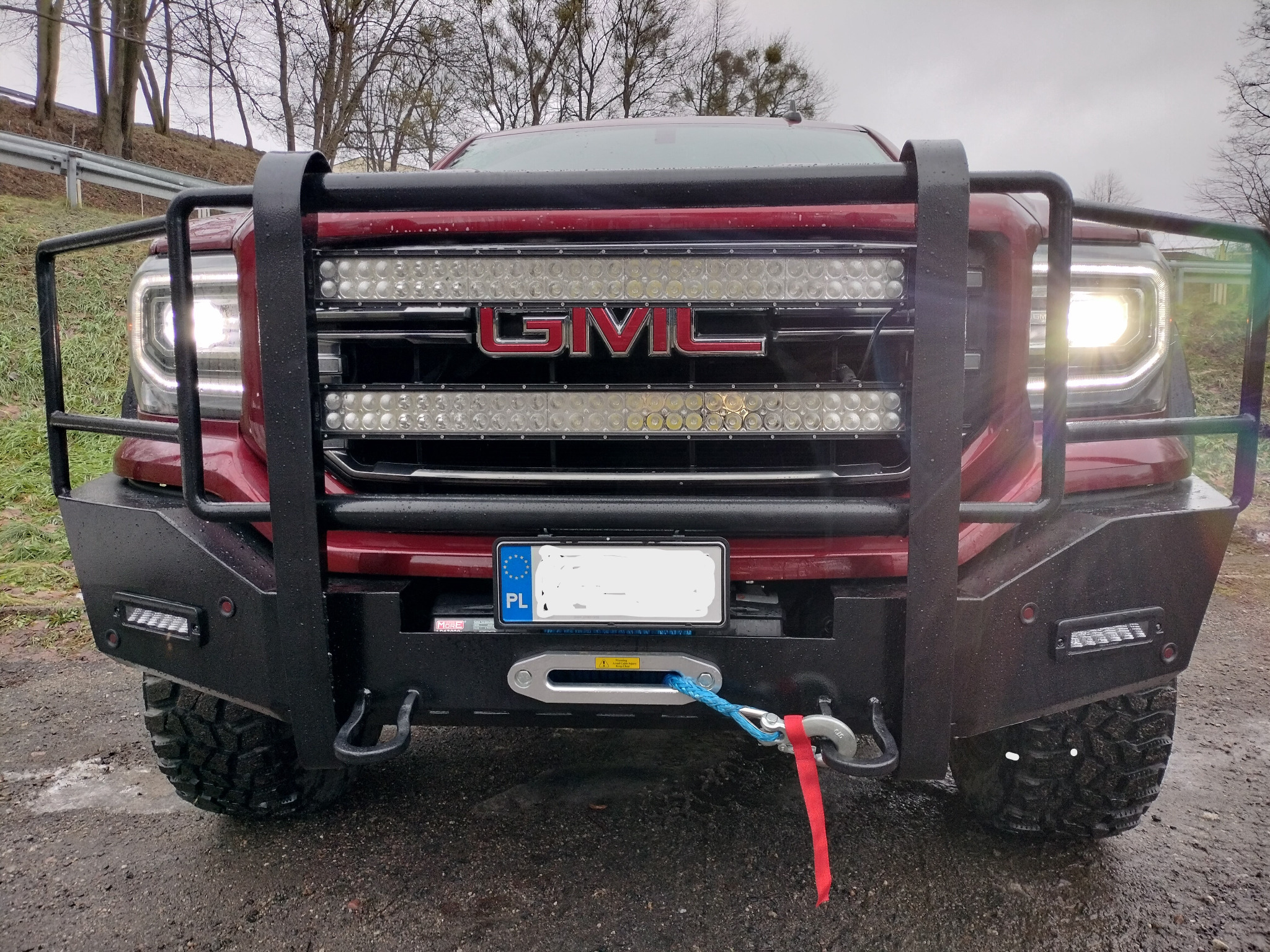 Przód czerwonego GMC Sierra z zamontowanym orurowaniem ochronnym, wyciągarką z liną syntetyczną i dodatkowym oświetleniem LED.