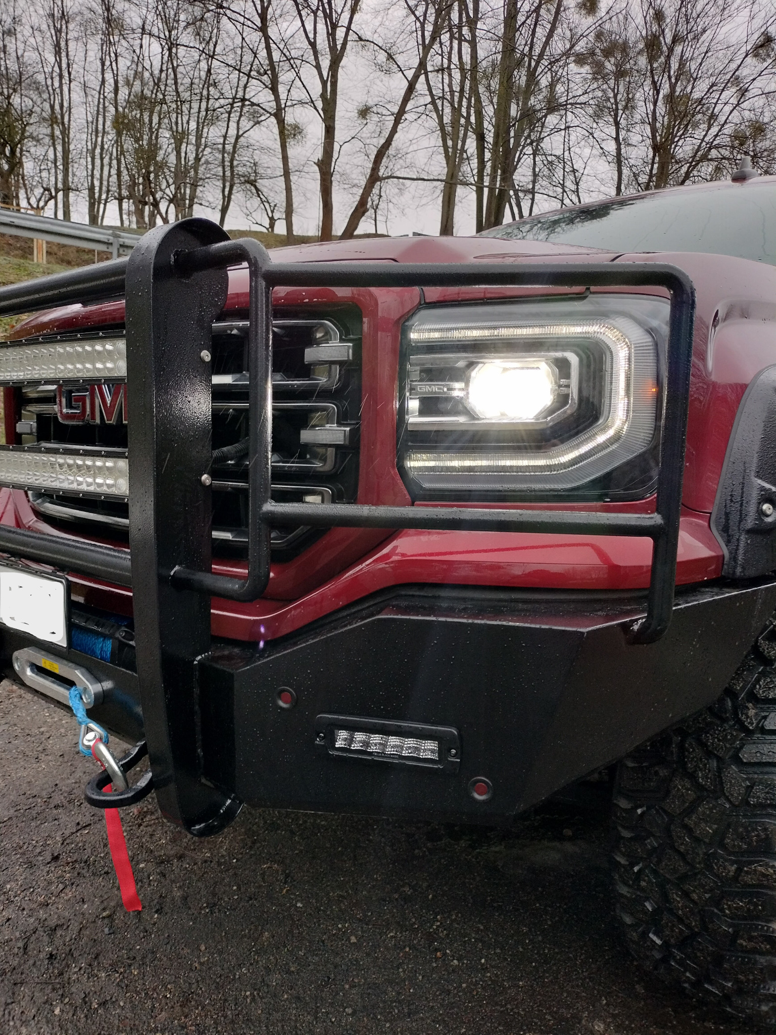 Czerwony samochód terenowy GMC z zamontowanym czarnym orurowaniem ochronnym i dodatkowymi światłami LED, widoczne detale opon terenowych i wyciągarki, na mokrym podłożu, drzewa w tle.