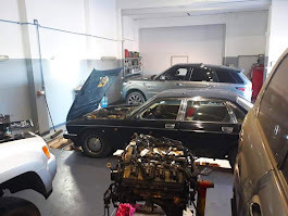 Wnętrze warsztatu samochodowego z podniesioną maską czarnego Jaguara, widoczny silnik na podłodze, w tle srebrny Range Rover i inne pojazdy.