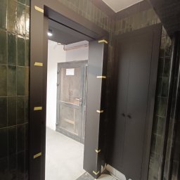 Rafał Nesterowicz - Ciemna, zabudowana szafa wnękowa z drzwiami na tle zielonych płytek, widoczna przez czarną ramę drzwiową zabezpieczoną taśmą malarską.