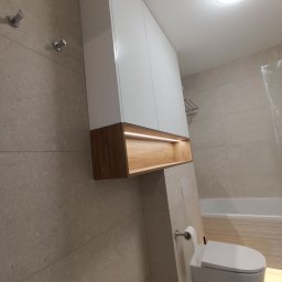 Rafał Nesterowicz - Biała szafka łazienkowa z drewnianym akcentem i podświetleniem LED nad toaletą podwieszaną w nowoczesnej łazience wykończonej beżowymi płytkami.