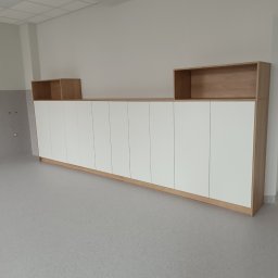 Rafał Nesterowicz - Długa szafa wnękowa z białymi frontami i drewnianymi elementami, zwieńczona otwartymi półkami, ustawiona w jasnym pomieszczeniu z szarą podłogą.