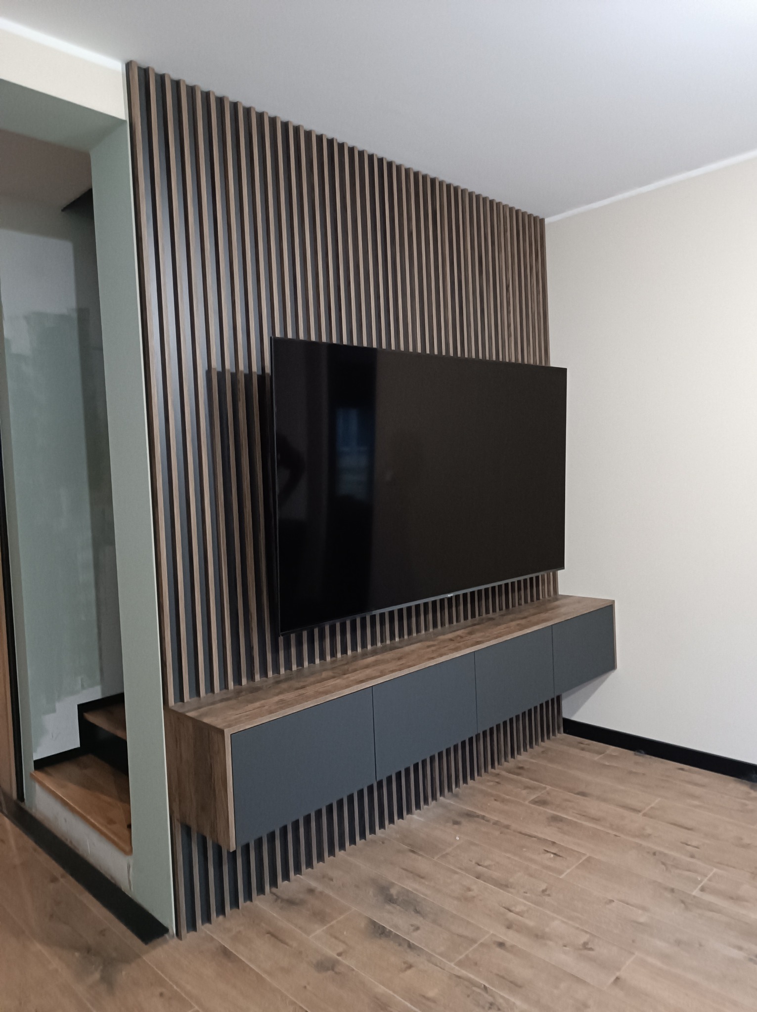 Nowoczesna ściana TV z drewnianymi lamelami, wisząca szafka RTV w odcieniach szarości i drewna, parkiet w jodełkę.