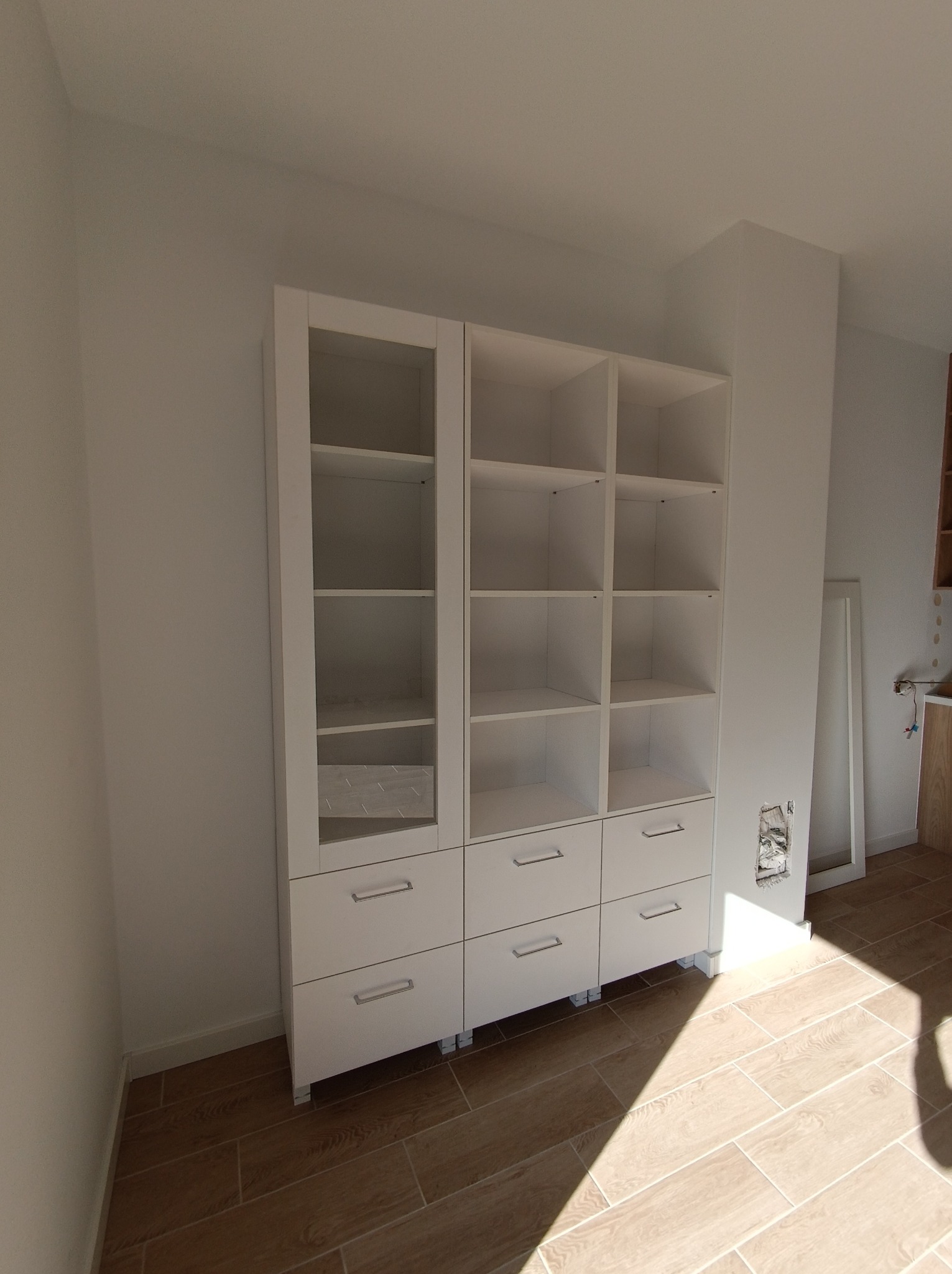 Biała szafa na wymiar z przeszkloną witryną i szufladami, widoczna na tle jasnych ścian i drewnopodobnej podłogi, z niedokończonymi pracami wykończeniowymi wokół gniazdka elektrycznego.