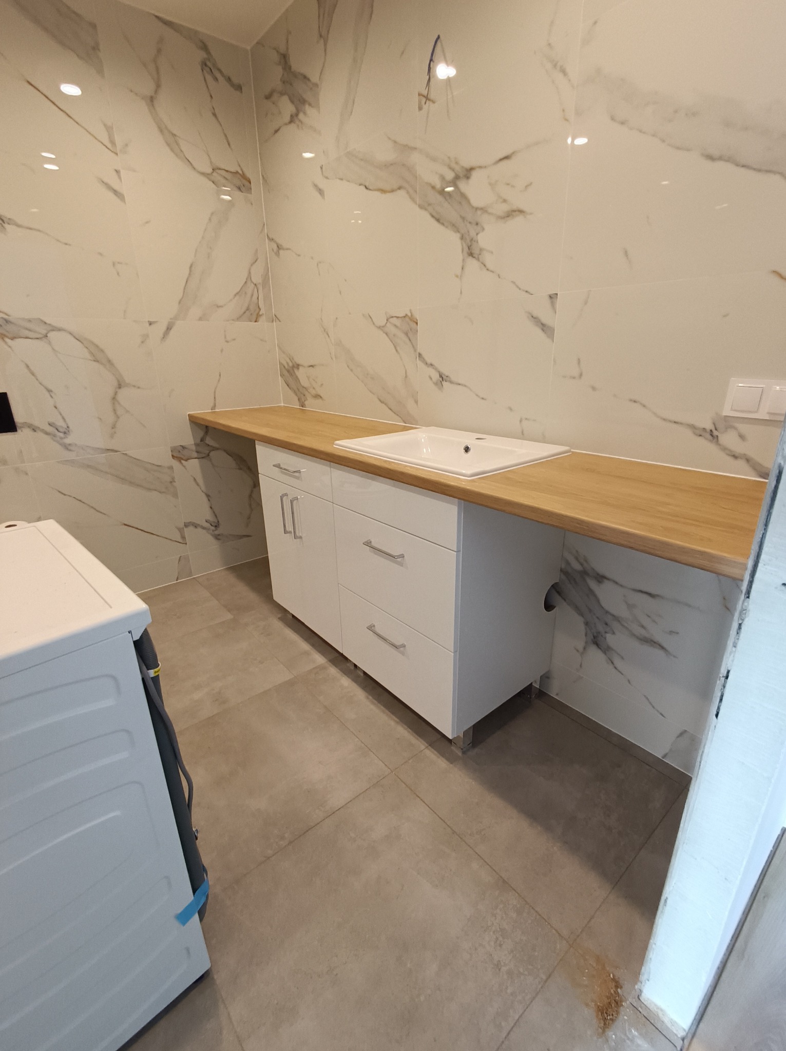 Biała szafka łazienkowa z trzema szufladami i szafką z blatem w kolorze jasnego drewna, umywalka wpuszczona w blat, marmurowe płytki na ścianie, widoczna pralka z boku.