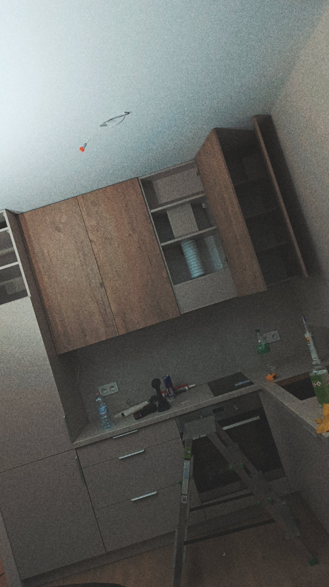 Montaż szarych szafek kuchennych z drewnianymi frontami na ścianie, widoczna drabina, butelka wody i narzędzia na blacie, surowy stan wykończenia.