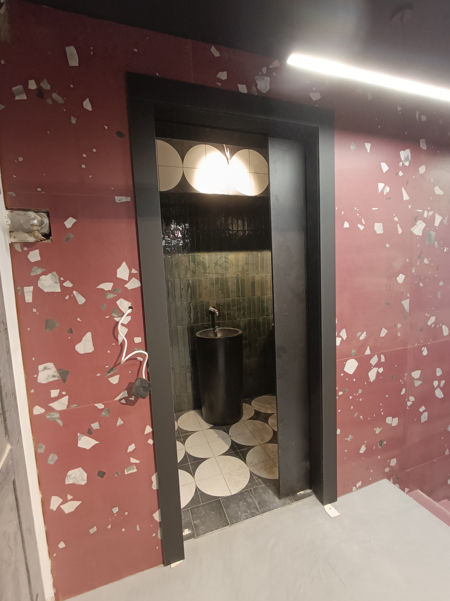 Nowoczesna łazienka w trakcie wykończenia: czerwona ściana z mozaiką, czarna umywalka kolumnowa, okrągłe płytki na podłodze, widoczne kable i niedokończone wykończenia.